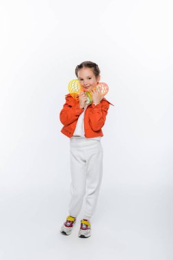 full length of happy kid in orange jacket playing with rainbow slinky on white clipart