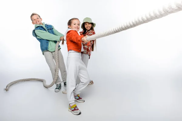 cheerful preteen children pulling rope while playing tug of war game on white