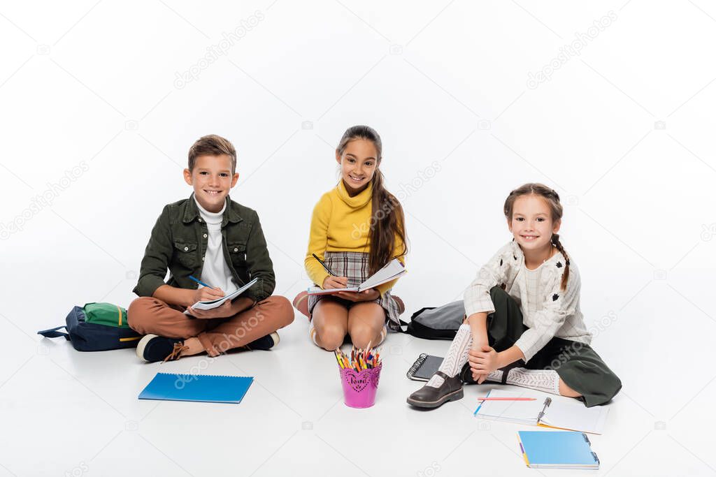 cheerful schoolboy and schoolgirls sitting near backpacks and notebooks while drawing on white 