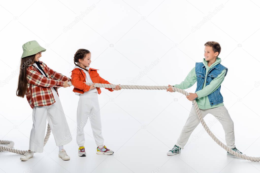 side view of cheerful girls pulling rope while playing tug of war game with boy on white