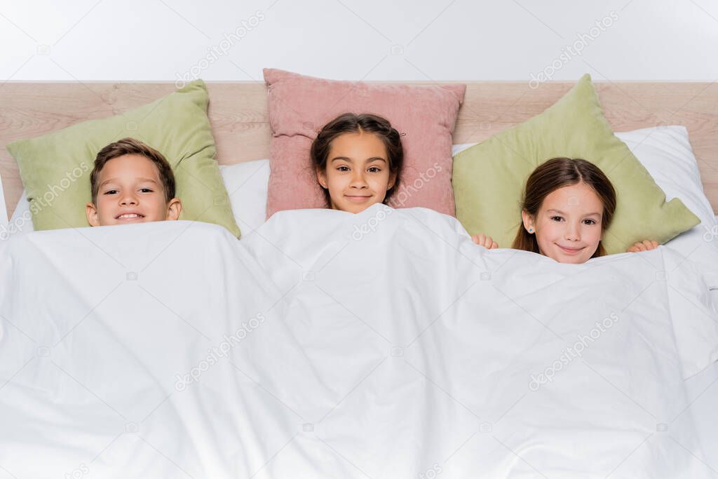 high angle view of cheerful kids lying under blanket isolated on white