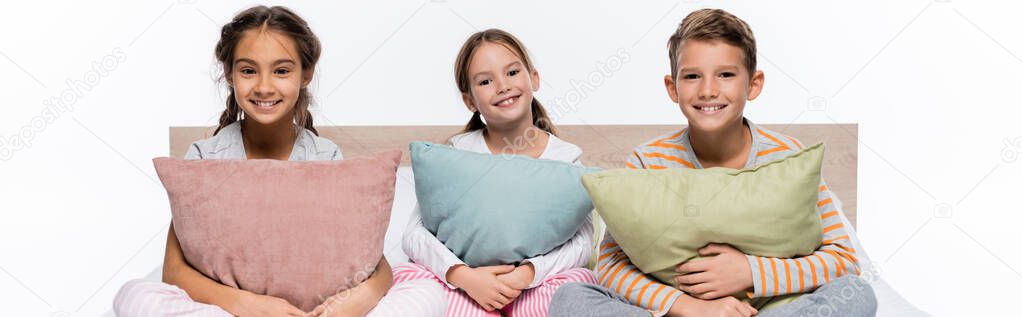joyful kids sitting on bed and hugging pillows isolated on white, banner
