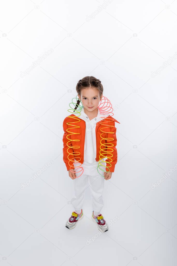 full length of smiling kid in orange jacket playing with rainbow slinky on white