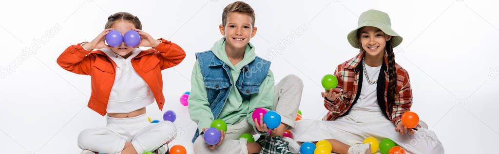 cheerful preteen kids sitting near friend covering eyes with colorful balls on white, banner