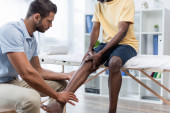 young rehabilitologist touching leg of african american man sitting on massage table