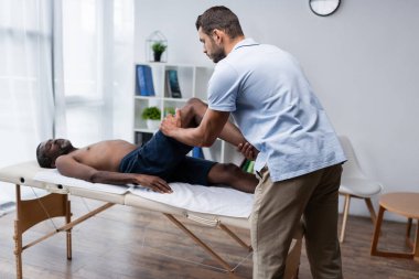 Genç kayropraktik uzmanı rehabilitasyon merkezinde yetişkin bir Afrikalı Amerikalı adamın bacağını esnetiyor.