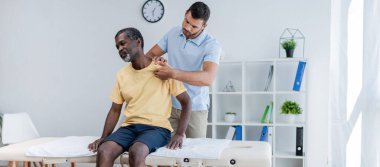 Orta yaşlı Afrikalı Amerikalı adam masaj masasında otururken rehabilitasyon uzmanı omzunu ve afişini inceliyor.