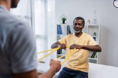Bulanık rehabilitasyon uzmanı ve Afrika kökenli Amerikalı hasta lastiklerle egzersiz yapıyor.