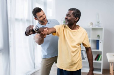 Genç antrenör mutlu Afrikalı Amerikalı bir adama rehabilitasyon merkezinde dambılla antrenman yaparken yardım ediyor.