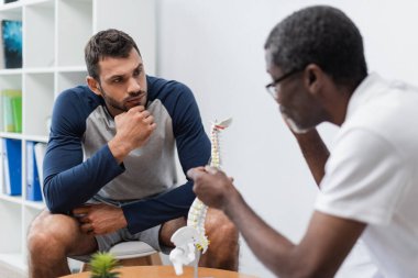 Bulanık Afrikalı Amerikalı doktor rehabilitasyon merkezinde ciddi bir adamın omurga modelini gösteriyor.