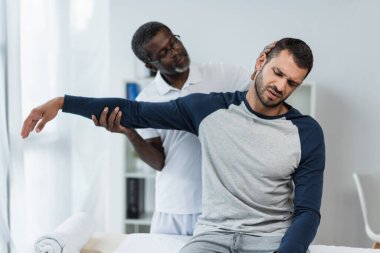 Olgun Afro-Amerikan rehabilitasyon uzmanı, çatık kaşlı ve acıdan muzdarip bir gencin kolunu tutuyor.
