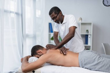 Olgun Afrikalı Amerikalı rehabilitasyon uzmanı genç bir adama sırt masajı yapıyor.