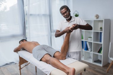 Afrikalı Amerikalı rehabilitasyon uzmanı klinikte genç bir adama masaj yapıyor.