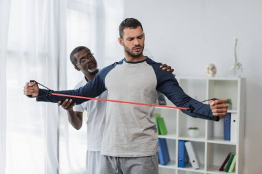 Afrikalı Amerikalı rehabilitasyon uzmanı direniş bandında genç adama yardım ediyor. 