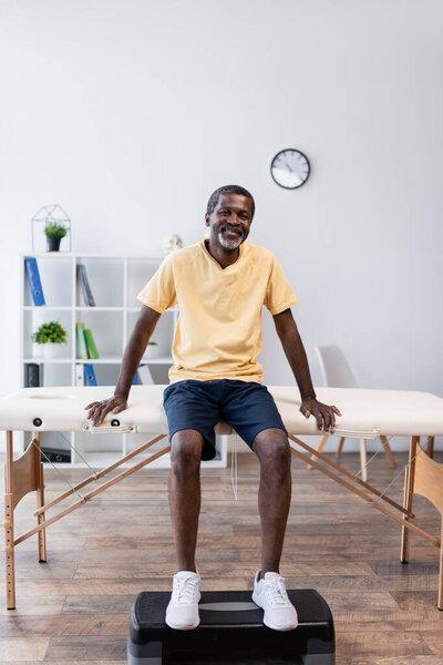 полный вид на Африканский мужчина среднего возраста, сидящий на массажном столе в реабилитационном центре