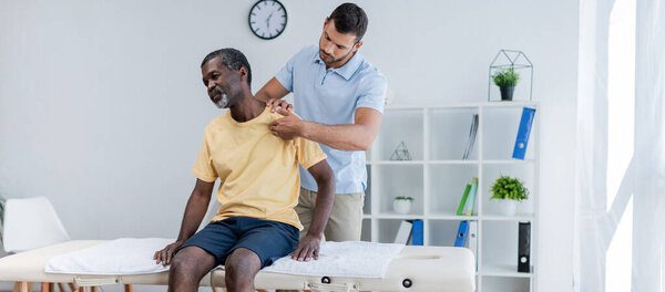 Африканский мужчина среднего возраста, сидящий на массажном столе, пока реабилитолог осматривает его плечо, баннер