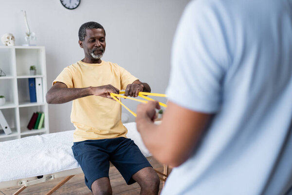 mature african american man doing exercise with elastics together with blurred rehabilitologist