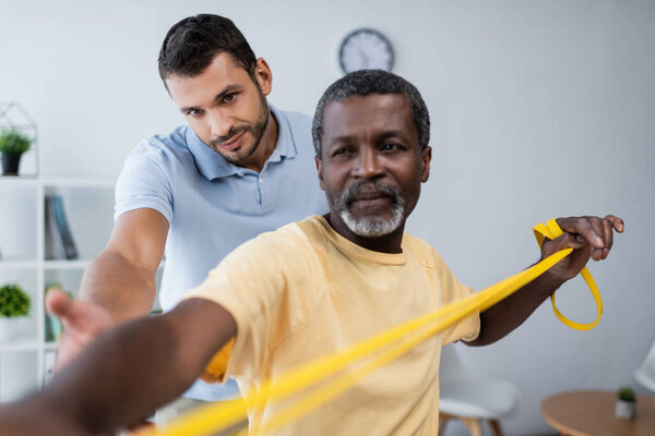 young instructor assisting african american man training with elastics in rehabilitation center
