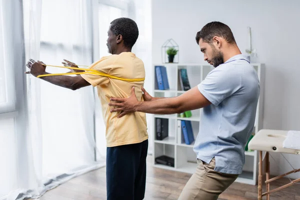 Young Trainer Supporting Mature African American Man Working Out Elastics — Stockfoto