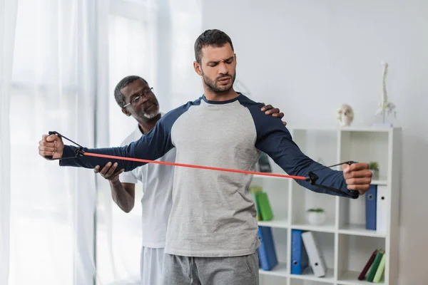 African American Rehabilitologist Assisting Young Man Training Resistance Band — Fotografia de Stock