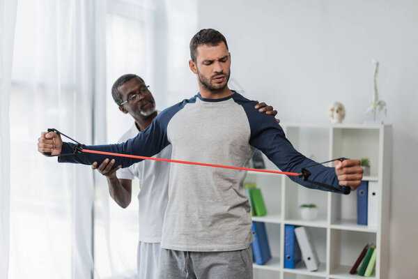 african american rehabilitologist assisting young man training with resistance band 