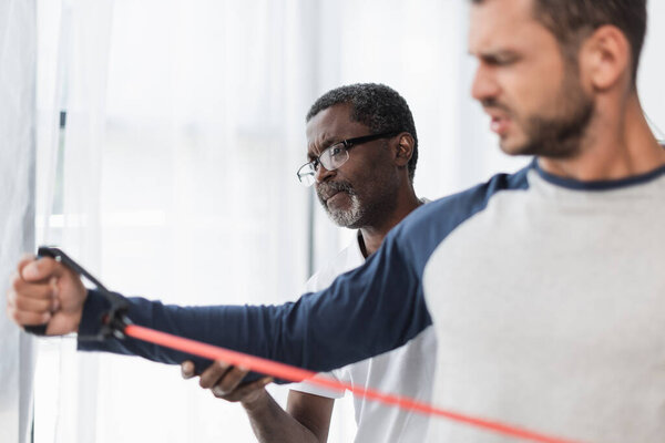 blurred man training with resistant band near mature african american physical therapist