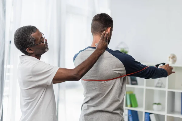 Afroameričtí Rehabilitační Trenér Pomáhá Muži Cvičit Odbojovou Kapelou — Stock fotografie