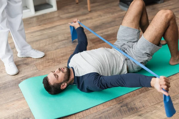 Ung Man Liggande Träningsmatta Och Träning Med Gummiband Nära Rehabilitolog — Stockfoto