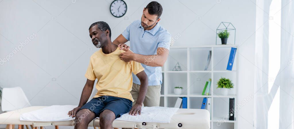 middle aged african american man sitting on massage table while rehabilitologist examining his shoulder, banner