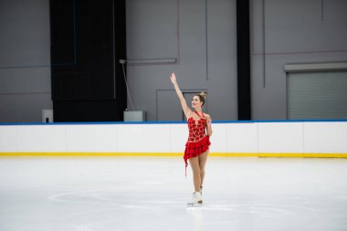 Kırmızı elbiseli neşeli artistik patinajcı elinde altın madalya ve buz arenasında el sallıyor.