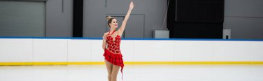 Kırmızı elbiseli neşeli artistik patinajcı elinde altın madalyayla buz arenasında el sallıyor.