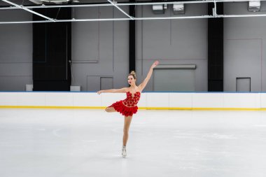 full length of figure skater in red dress performing camel spin in ice arena clipart