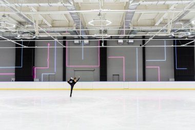 back view of young woman in black bodysuit skating in professional ice arena clipart