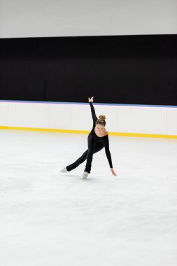 full length of young professional figure skater in black bodysuit skating with outstretched hand in ice arena  clipart