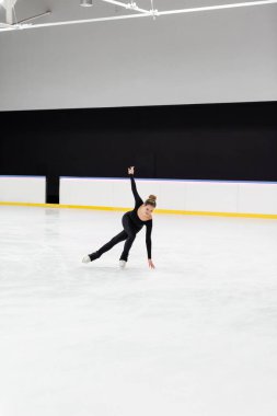 Arenada uzanmış ellerle buz pateni yapan siyah elbiseli genç artistik patinajcı. 