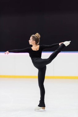 side view of professional figure skater in black bodysuit stretching with outstretched hand in ice arena clipart