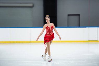 full length of cheerful woman in red dress figure skating on professional ice rink clipart