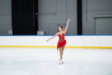 full length of happy woman in red dress figure skating in professional ice rink clipart