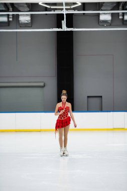 Kırmızı elbiseli genç artistik patinajcı buz pistinde altın madalyaya bakıyor.