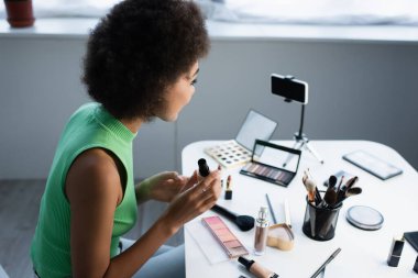 Afro-Amerikan blog yazarının kozmetik ve akıllı telefonun yanında yüz temeli tutarken yan görüntüsü 