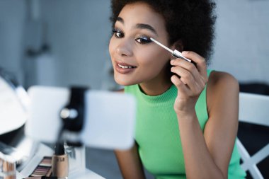 Positive african american blogger looking at smartphone while applying eye shadows  clipart