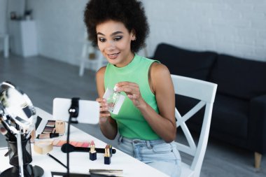 Neşeli Afro-Amerikan blog yazarı kozmetik ve akıllı telefonun yanında parfüm tutuyor. 