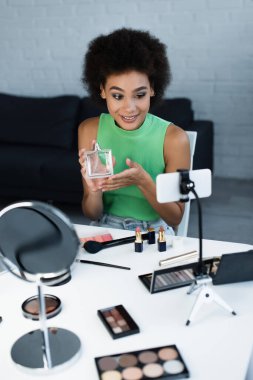 Afro-Amerikan blog yazarı akıllı telefon, ayna ve dekoratif makyaj malzemelerinin yanında parfüm tutuyor. 