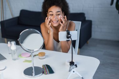 Neşeli Afro-Amerikan blogcu cep telefonu ve evde yeşim taşıma aracının yanında kozmetik krem uyguluyor. 