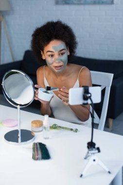 Evde akıllı telefonun yanında kilden maske ve kozmetik fırça tutan Afrikalı Amerikalı blogcu 
