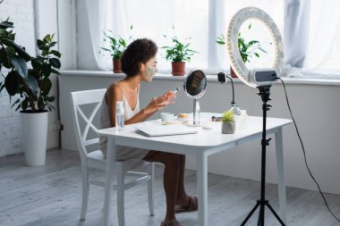 Yüzünde kil maske olan Afro-Amerikan blog yazarı akıllı telefonun yanında kozmetik fırça tutuyor. 