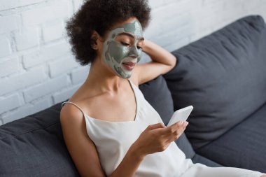 Yüzünde kil maske olan gülümseyen Afro-Amerikalı kadın evdeki koltukta cep telefonu kullanıyor. 