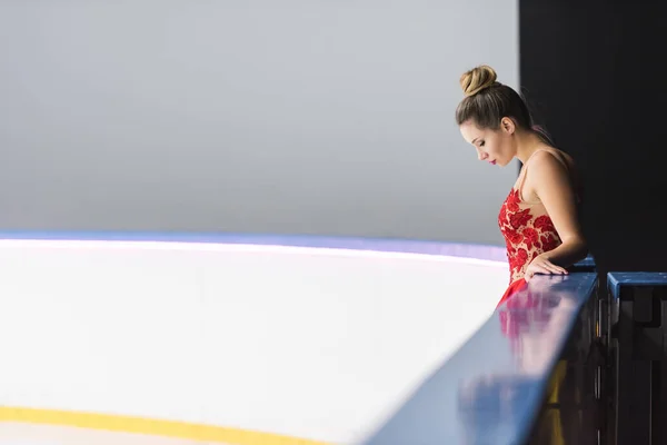 アイスリンクに立つ赤いドレス姿の若いフィギュアスケート選手のサイドビュー — ストック写真