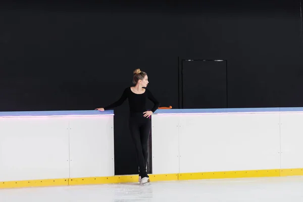 黒のボディスーツ姿の若いフィギュアスケート選手が凍った氷のアリーナ近くの腰に手を置き — ストック写真