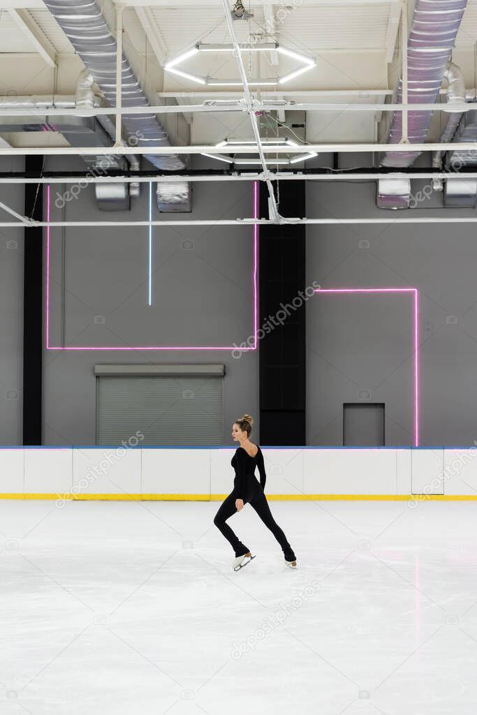 full length of young woman in black bodysuit skating in professional ice arena